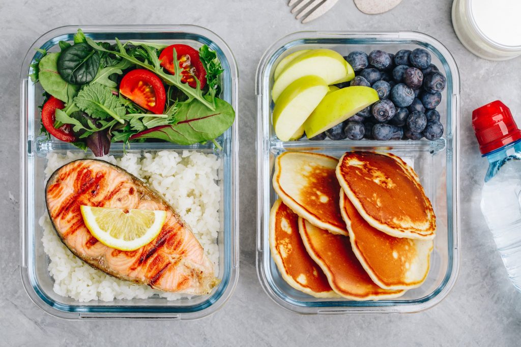 Meal prep containers with salmon, rice, green salad and pancakes, apple, blueberry.