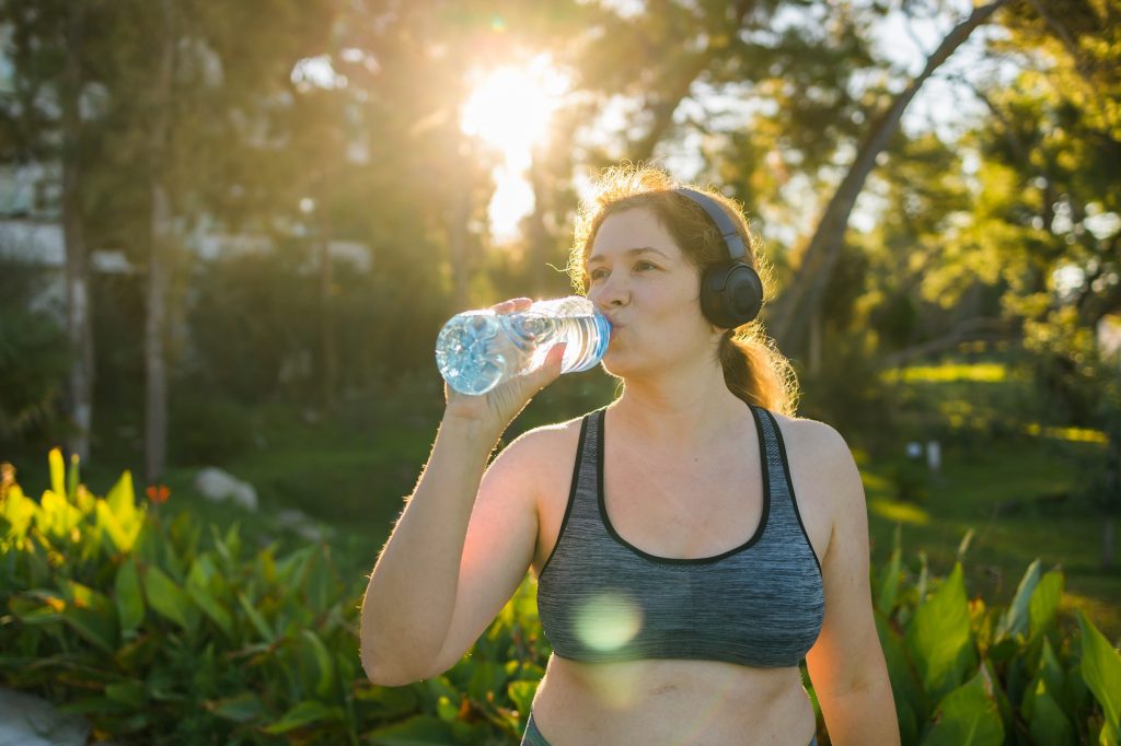 hydration in health, Overweight woman drinking water after jogging in the park. Portrait of young plus-size thirsty woman